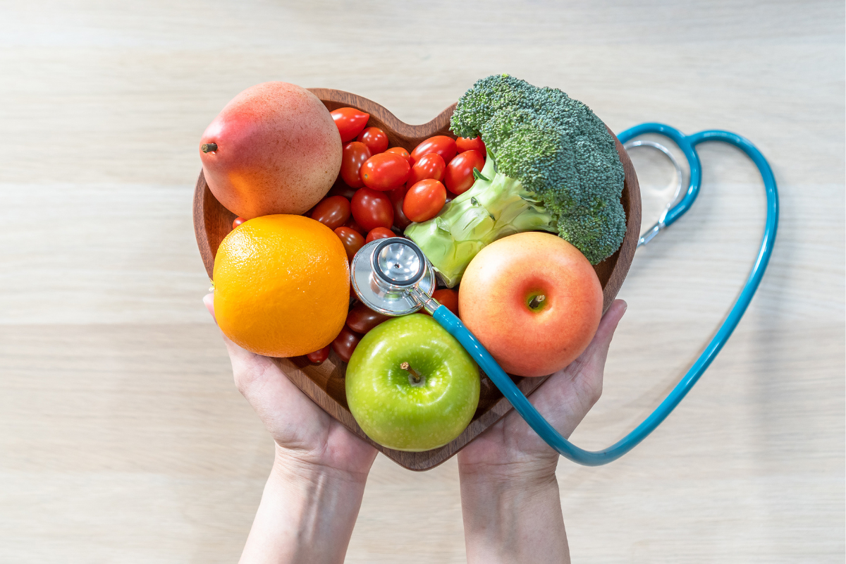 Hands holding a heart bowl with stethoscope, fruits, and broccoli